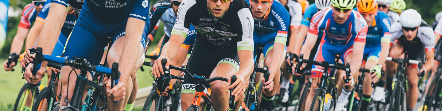 Groupe de coureurs cyclistes serrés en peloton sur une route, portant des maillots colorés dont un cycliste au centre en tenue noire et blanche arborant le logo 'NEAT' et des sportifs en bleu avec l'inscription 'MALOJA PUSH BIKES'.