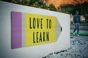 Vue d’une pancarte extérieure en forme de crayon fixée à un mur blanc, affichant en lettres noires sur fond jaune le texte exact « LOVE TO LEARN ». À gauche, la pointe et la gomme du crayon sont colorées en beige et mauve. Un homme en sweat à capuche et lunettes de soleil marche en arrière-plan sur un trottoir bordé de galets blancs.