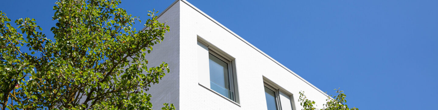 Façade d'un bâtiment résidentiel blanc à lignes épurées avec fenêtres carrées, entouré d'arbres verts sous un ciel bleu clair