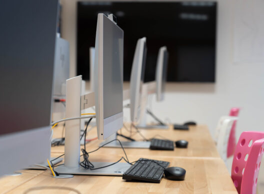 Vue latérale d’une rangée d’ordinateurs de bureau HP tout-en-un alignés sur une table en bois avec claviers et souris, chaise rose au premier plan