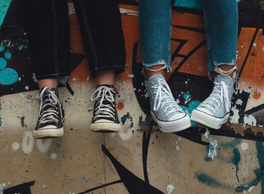 Vue rapprochée de deux paires de baskets montantes – une paire noire sur jean noir et une paire grise sur jean bleu déchiré – les pieds pendant devant un mur de béton recouvert de graffitis orange, turquoise et noir.