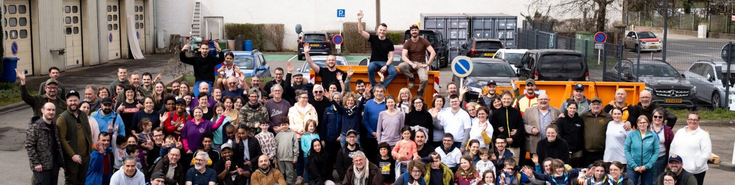 Photo d’une grande trentaine de bénévoles – adultes, enfants et scouts – rassemblés sur un parking urbain devant des portes de garage. Tous sont souriants, certains portent des chasubles haute visibilité, un t-shirt «MOIEN», et saluent de la main. Au centre, un chien. À l’arrière-plan, bennes et voitures garées.